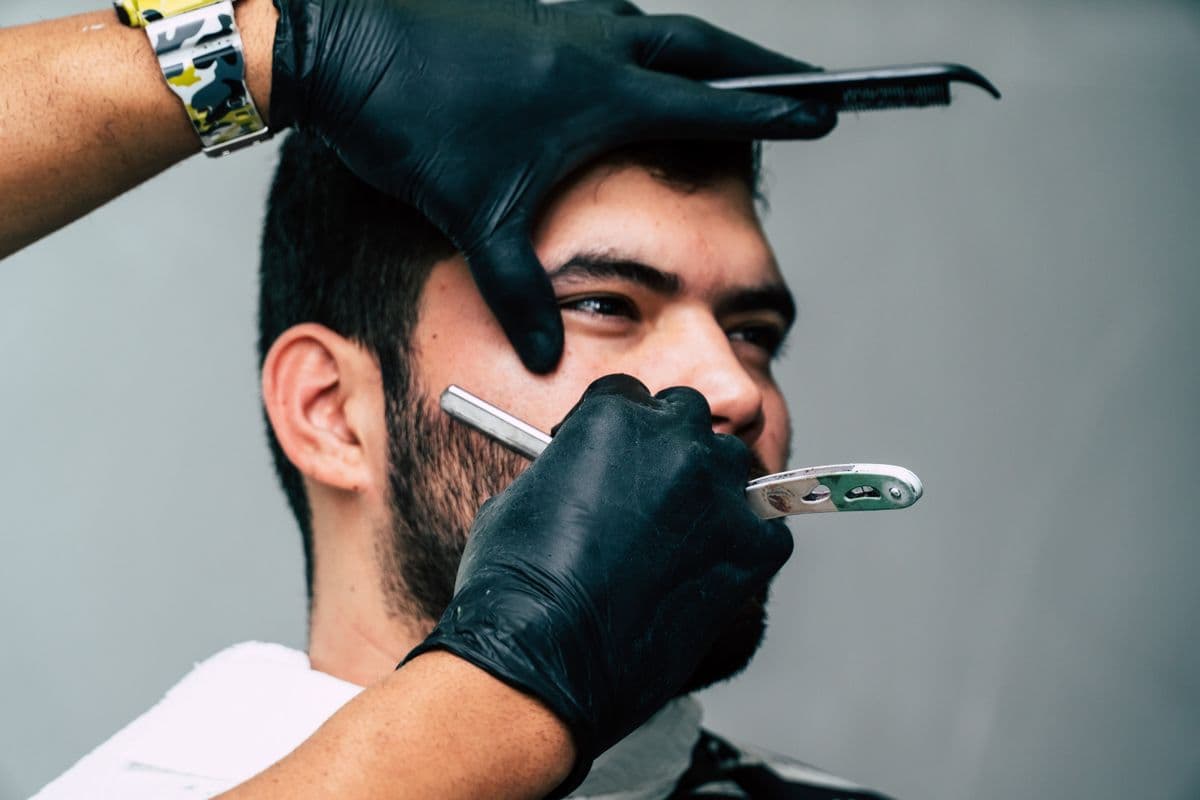 Un barbero con guantes negros afeitando con navaja la barba de un cliente.