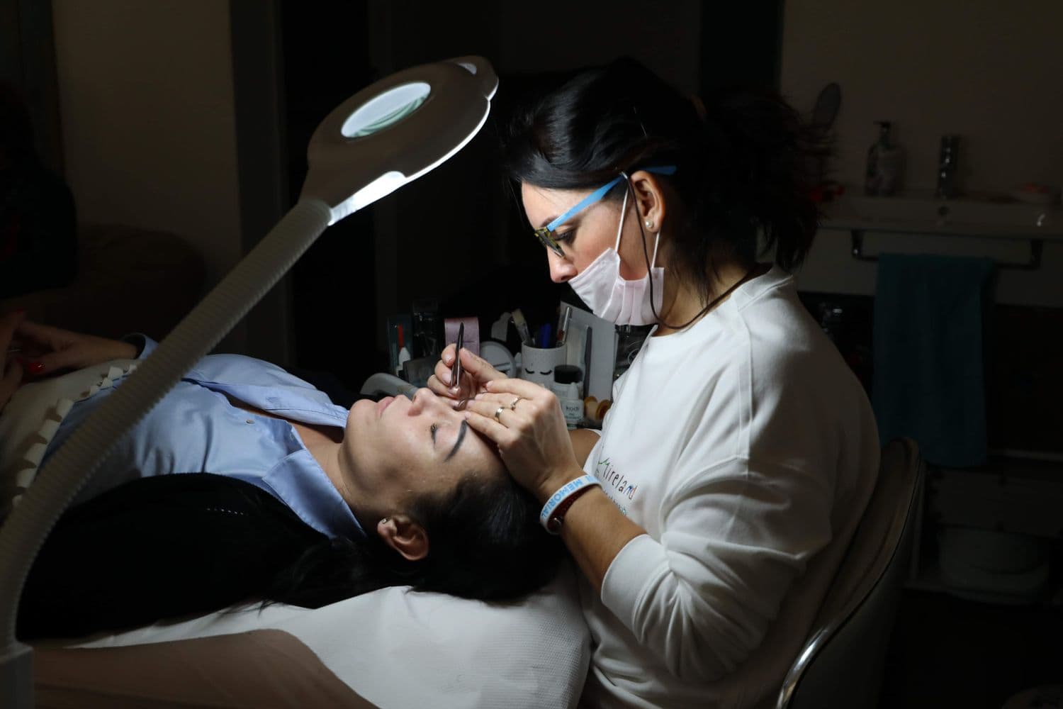 Una especialista diseñando las cejas de su clienta, recostada bajo una lámpara de aumento.