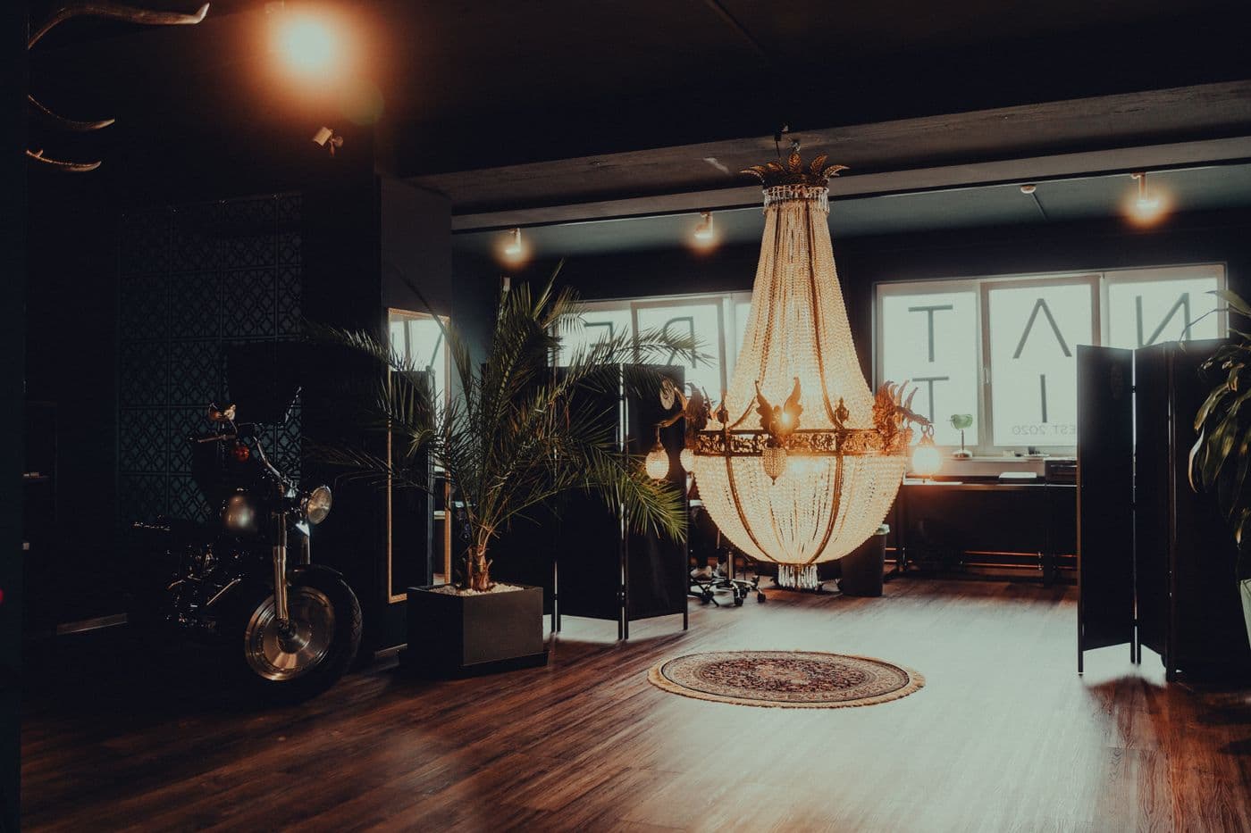 El interior de un estudio de tatuajes con piso de madera, una lámpara de araña, plantas y una moto al fondo.